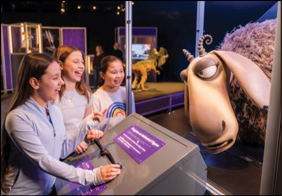 Three children smiling and interacting with a museum exhibit featuring a large animated animal figure. Description generated by AI