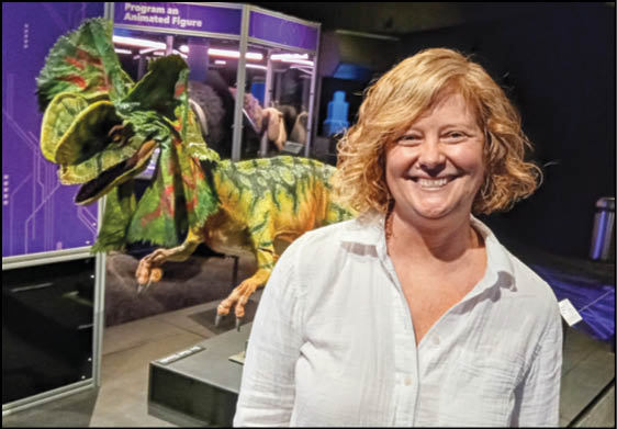 A person with curly hair wearing a white shirt is smiling and standing next to a colorful dinosaur model in a museum or exhibit setting. Description generated by AI