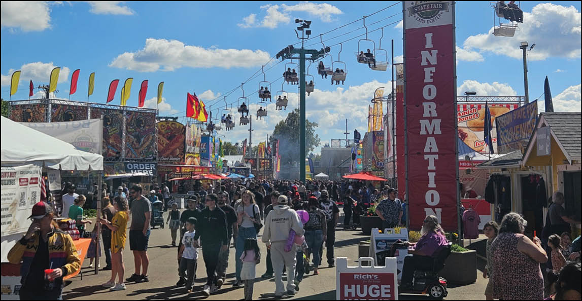 A crowded outdoor fair with people walking along a pathway lined with food stalls, colorful flags, and a chairlift overhead under a partly cloudy sky. Description generated by AI