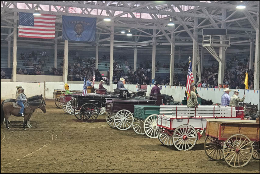 Several people dressed in western attire are riding horses and horse-drawn wagons inside an indoor arena decorated with American and state flags. Description generated by AI