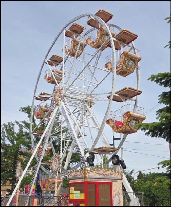 A vintage-style Ferris wheel with ornate, open-air seats is set up outdoors against a clear sky with trees and a small ticket booth at its base. Description generated by AI