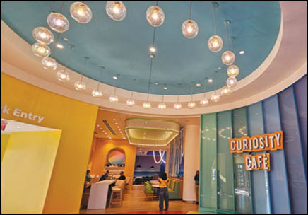 A brightly lit caf interior with a circular chandelier, colorful walls, and people sitting and standing near tables under a sign that reads “Curiosity Caf ."  Description generated by AI