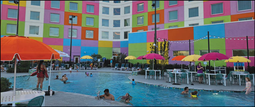 People are swimming and relaxing around a pool with colorful umbrellas and a vibrant, multicolored building in the background. Description generated by AI