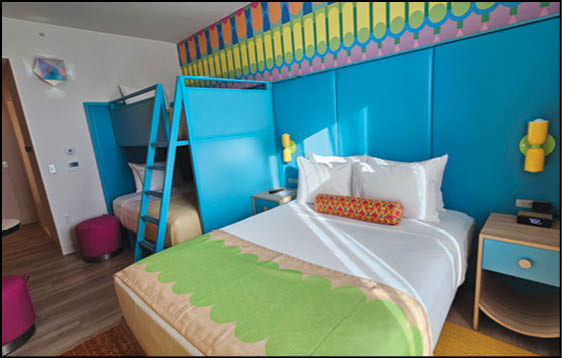 A brightly colored hotel room features a large bed with white linens and a green blanket, a bunk bed with a ladder, vibrant blue padded walls, and playful yellow and pink wall decorations. Description generated by AI