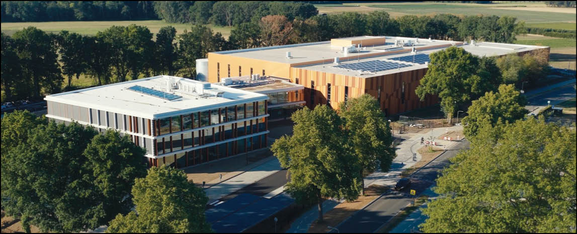 Aerial view of a modern campus with two large buildings surrounded by trees and adjacent to a road. Description generated by AI