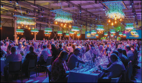 A large group of people seated at long tables in a dimly lit banquet hall with decorative hanging lights and screens displaying the word “VERGOS." Description generated by AI
