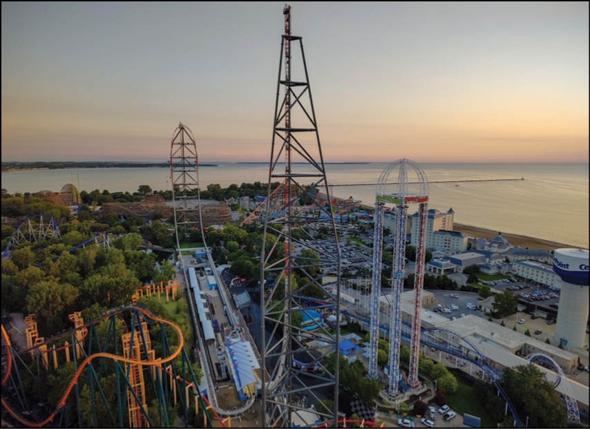Aerial view of an amusement park at sunset featuring roller coasters, tall thrill rides, a parking lot, and a body of water in the background. Description generated by AI