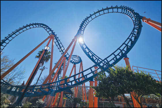 A roller coaster with orange supports and blue tracks features a large loop against a clear blue sky with sunlight shining through. Description generated by AI