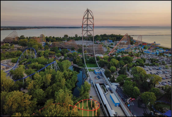 An amusement park at sunset featuring multiple roller coasters, lush green trees, a body of water, and a parking lot. Description generated by AI
