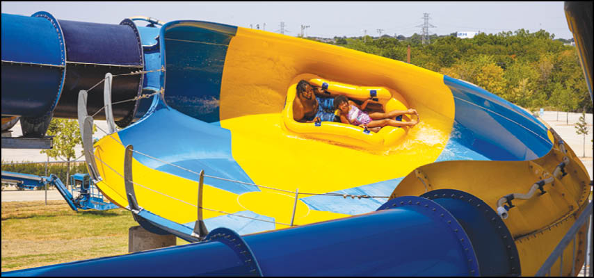 Two people wearing casual clothing are riding together on a yellow raft inside a large blue and yellow water slide at a water park. Description generated by AI