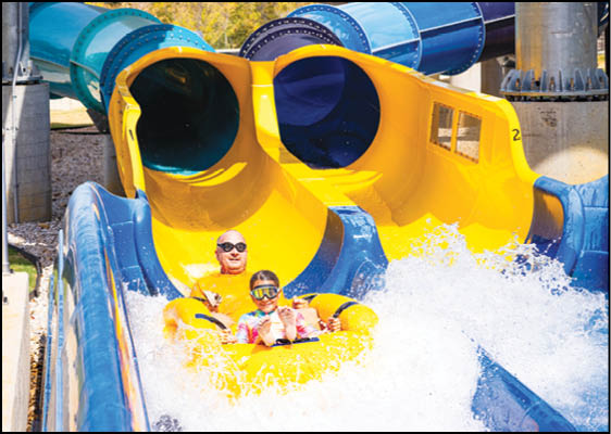 Two people wearing goggles are riding together on a yellow inflatable raft coming out of parallel blue and yellow water slides with splashing water around them. Description generated by AI