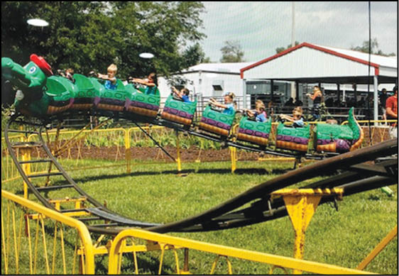 Children are riding a small green dragon-themed roller coaster at an outdoor amusement park with yellow safety railings and a building in the background. Description generated by AI