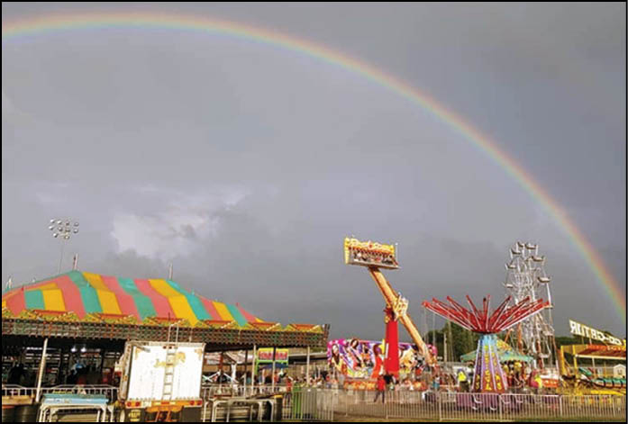 A colorful carnival scene with various rides and a large rainbow arching across a cloudy sky. Description generated by AI