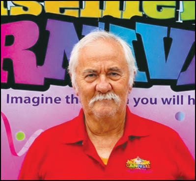 A senior person with white hair and a mustache wearing a red polo shirt with a colorful logo stands in front of a vibrant background with large multicolored letters and partial text. Description generated by AI