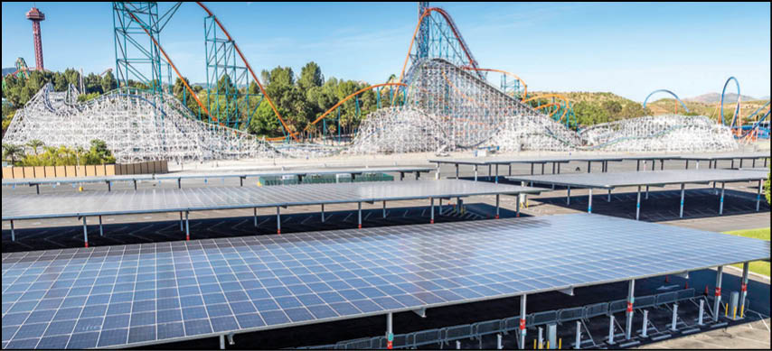 A large solar panel installation is in the foreground with multiple roller coasters and trees visible in the background under a clear blue sky. Description generated by AI