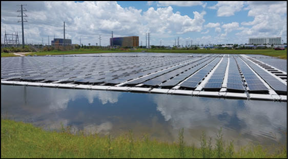 A large array of solar panels is installed on a floating platform on a body of water with grassy banks and industrial buildings in the background under a partly cloudy sky. Description generated by AI