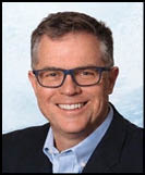 A person with short hair and glasses is smiling while wearing a dark blazer over a light blue collared shirt against a light background. Description generated by AI