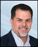 A person with short dark hair and a beard is smiling and wearing a dark blazer over a light checkered shirt against a light blue background. Description generated by AI