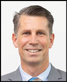 A person with neatly combed short hair is smiling and wearing a gray suit jacket, light blue shirt, and striped tie against a plain white background. Description generated by AI