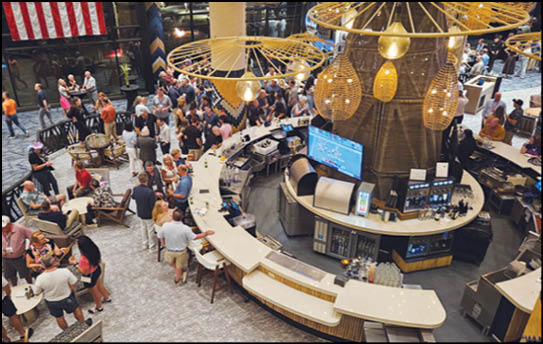 A large group of people are gathered around and sitting near a circular bar in a spacious indoor venue with modern lighting and a large screen. Description generated by AI