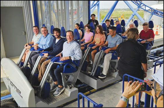 A group of people seated and smiling on a roller coaster ride at an amusement park with an attendant nearby and a person taking a photo. Description generated by AI