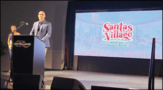 A person in a suit is speaking at a podium on a stage with a large screen behind them displaying the logo and slogan for Santa's Village. Description generated by AI