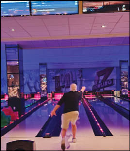 A person wearing a black shirt and light shorts is bowling in a brightly lit bowling alley with colorful lane lights and screens overhead. Description generated by AI