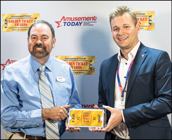 Two adults dressed in business attire smile while holding a framed Golden Ticket Award at an event with an Amusement Today backdrop. Description generated by AI