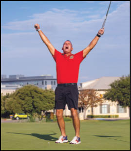 A person wearing a red shirt and black shorts is standing on a golf course with arms raised in celebration while holding a golf club. Description generated by AI