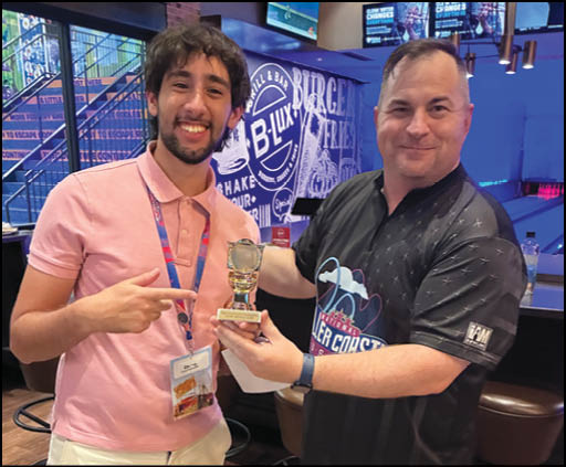 Two adults smiling indoors with one holding a small trophy and the other pointing at it. Description generated by AI