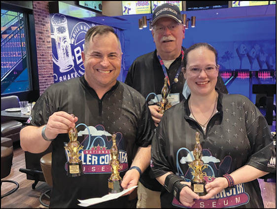 Three adults wearing matching shirts smile and hold trophies in a bowling alley setting. Description generated by AI