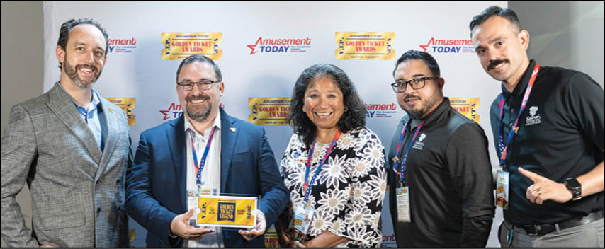 Five adults wearing conference badges stand smiling in front of a backdrop with logos while one holds a yellow award box. Description generated by AI