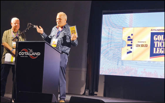Two adults stand on a stage at a podium with a microphone, one holding a book and speaking, while a large screen displays a VIP ticket graphic in the background. Description generated by AI