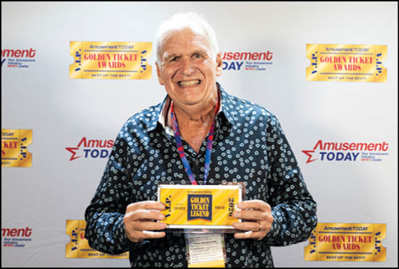 A senior with white hair wearing a patterned dark shirt smiles while holding a Golden Ticket Legend award in front of a backdrop featuring Amusement Today and Golden Ticket Awards logos. Description generated by AI
