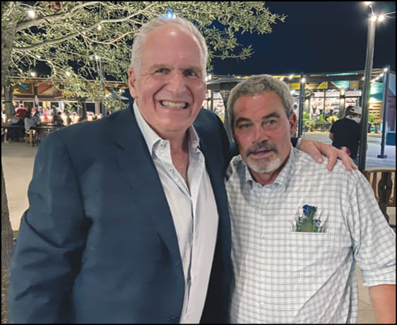 Two adults are smiling and posing closely together outdoors at night with lights and trees in the background. Description generated by AI