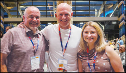 Three adults smiling and standing close together indoors, each wearing a lanyard with badges, with a lively background of other people and modern decor. Description generated by AI