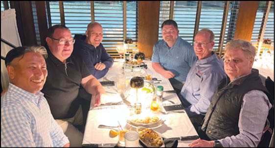 Six adults sitting around a well-lit dining table with plates of food and drinks, smiling and engaging in conversation in a cozy indoor setting. Description generated by AI