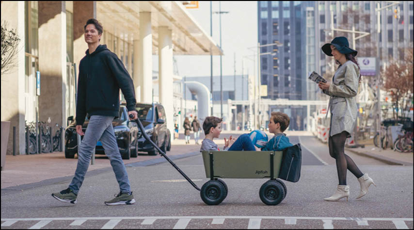 A person in casual clothing pulls a wagon carrying two children while another person in a hat and light-colored outfit walks nearby reading a book on a city street. Description generated by AI