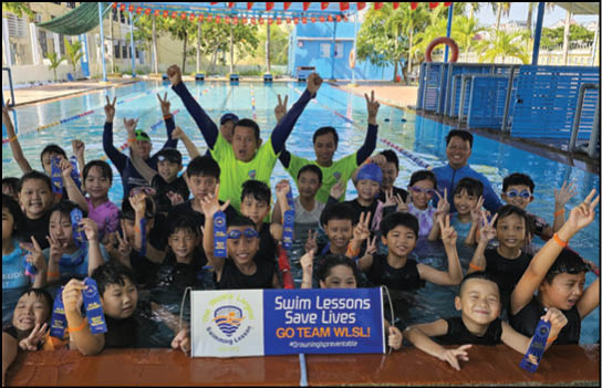 A group of people in a pool with a sign that says “Swim Lessons Save Lives". Description generated by AI