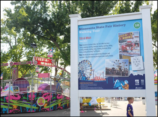 A colorful Tilt-A-Whirl amusement ride is next to a large informational sign about the Minnesota State Fair history walking tour with images of the ride and a child walking nearby. Description generated by AI