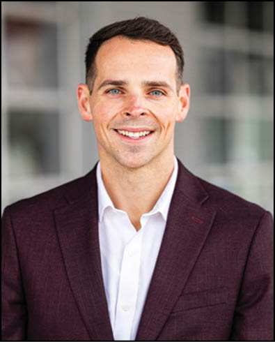 A person with short dark hair wearing a dark maroon blazer and white shirt is smiling against a blurred indoor background. Description generated by AI