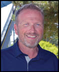 A smiling adult with short hair and a beard wearing a navy blue collared shirt stands outdoors near trees and a metal structure. Description generated by AI