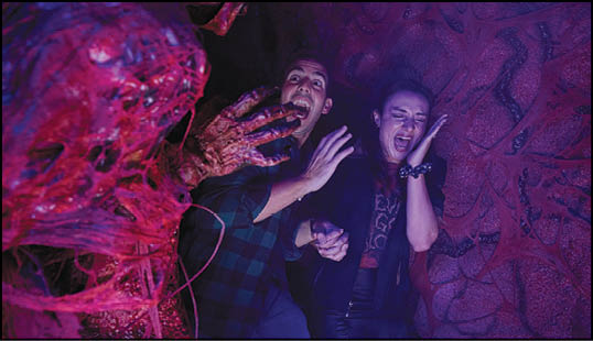 Two adults holding hands and reacting with fear as a grotesque red creature reaches toward them in a dark, eerie setting. Description generated by AI