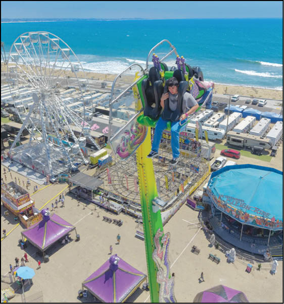 A person wearing a helmet and harness is suspended high above a beachside amusement park with a Ferris wheel, tents, and a carousel visible below. Description generated by AI