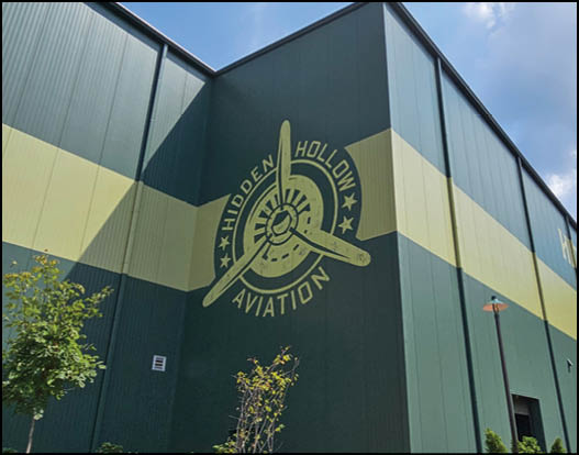 A large green and yellow building corner features a painted logo of a propeller with the words Hidden Hollow Aviation surrounded by stars. Description generated by AI