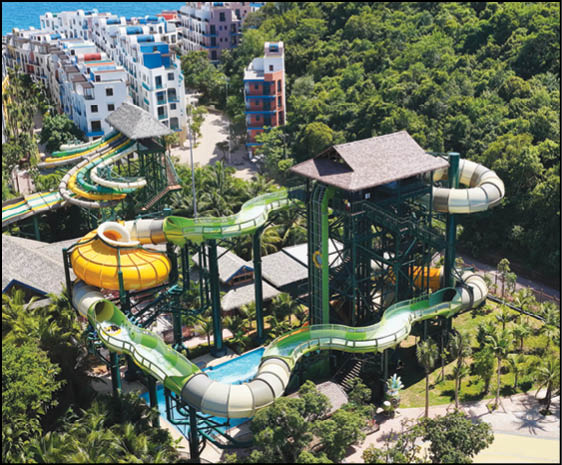 A large outdoor water park features multiple twisting water slides supported by green metal structures surrounded by lush greenery and nearby buildings. Description generated by AI