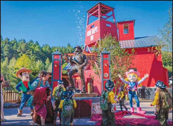 Adults and children dressed in firefighter gear and casual clothing gather around a bronze firefighter statue in front of a red fire department building with a tower. Description generated by AI