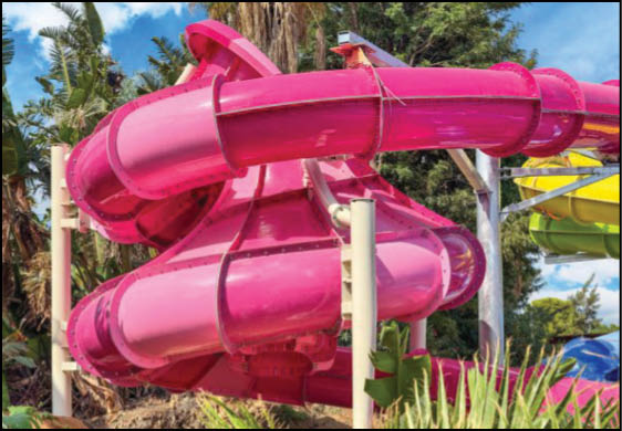 A bright pink spiral water slide is set outdoors surrounded by green plants and trees under a blue sky. Description generated by AI