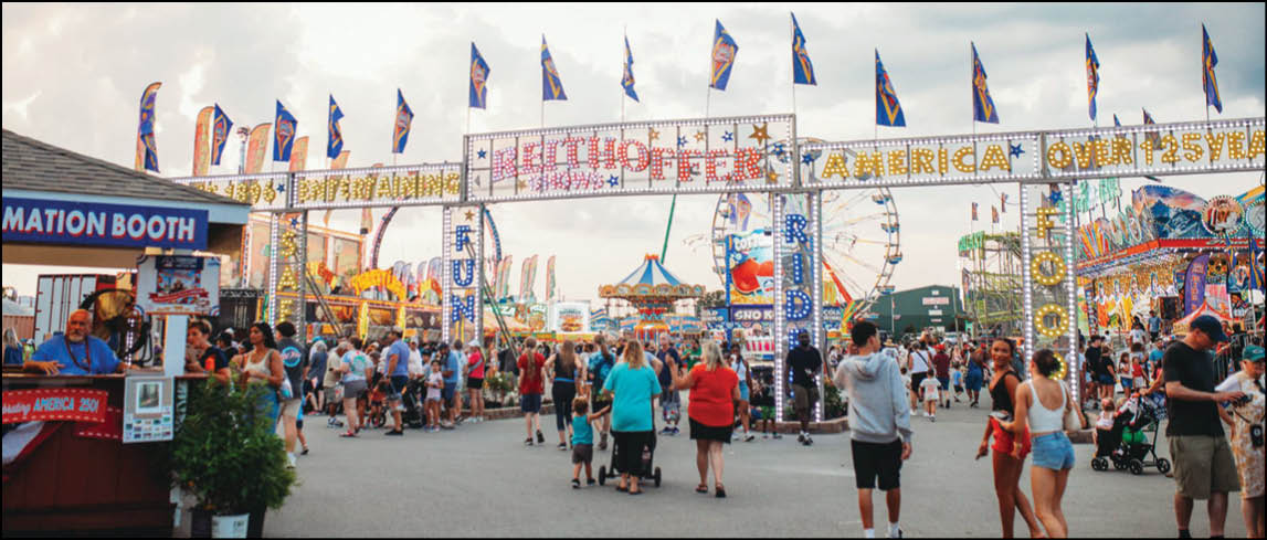 A crowded outdoor fair with people walking and standing near colorful booths, flags, and amusement rides under a partly cloudy sky. Description generated by AI