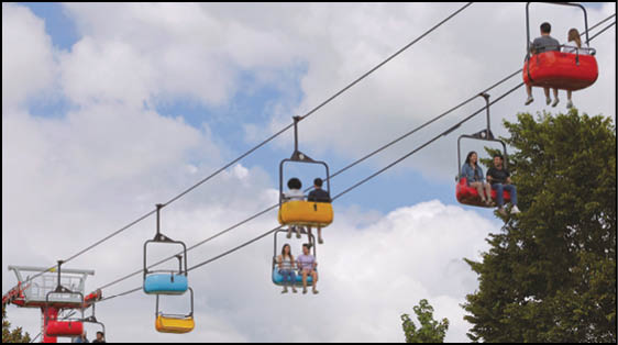 People are riding colorful open-air chairlifts suspended on cables against a partly cloudy sky with trees below. Description generated by AI